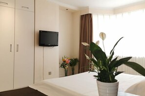 Habitación de hotel con cama, televisor montado en la pared, planta en maceta y ventana con cortinas.