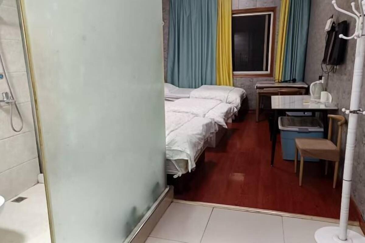Una habitación con dos camas, una mesita, una silla, un bote de basura y un ventanal con cortinas.