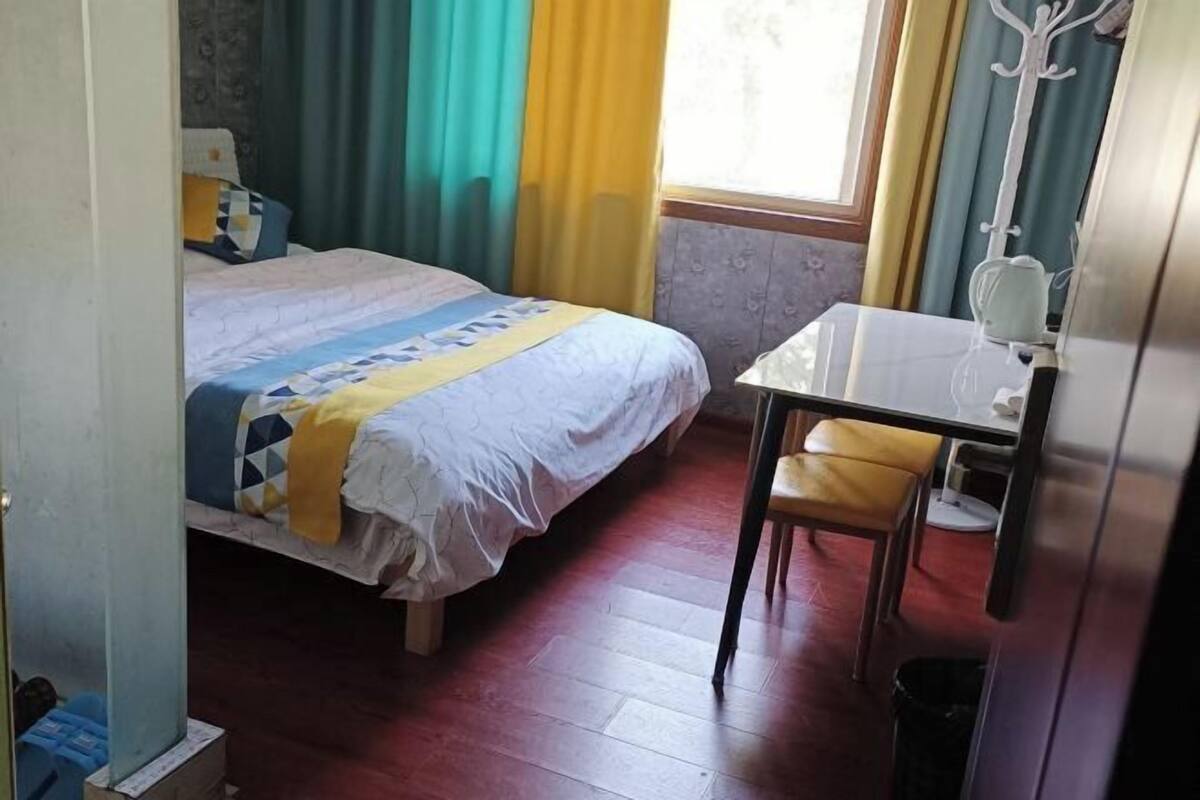 Una habitación con cama, una mesita, una silla, un espejo y un ventanal con cortinas.