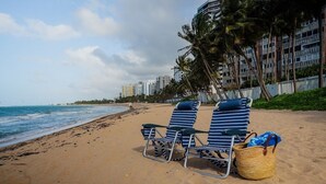 A beach with two striped lounge chairs and a woven basket on the sand, overlooking the ocean and a row of buildings.