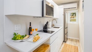 A compact kitchen with white cabinetry, a built-in microwave, and a sink. There is a small wooden bench, a bowl of fruit, and a bottle of wine on the counter.