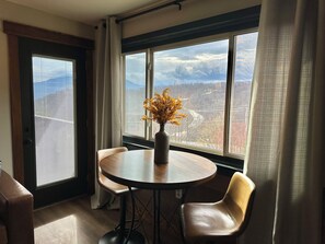 Zona pranzo con tavolo rotondo, due sedie, un vaso con fiori gialli e vista sulle montagne attraverso ampie finestre.