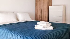 Een slaapkamer met een bed, witte kussens, een blauw geweven sprei, een houten accentmuur en een witte dressoir.