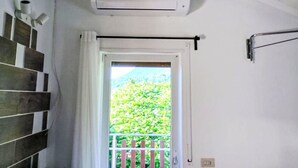 Een kamer met een witte airconditioner aan de muur, een houten trap met een wit gordijn, en een openslaande deur naar een groene tuin.