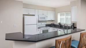 Una cocina compacta con armarios blancos, encimera negra, microondas, estufa y lavaplatos. Hay dos sillas de madera con cojines azules.