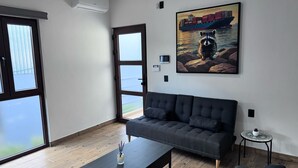 A modern living room with a dark grey sofa, a wooden coffee table, and a small side table. There is a large window with a view of the outside, and a painting of a raccoon on rocks is on the wall.
