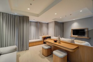 Une chambre d’hôtel moderne, équipée d’un canapé, d’une petite table avec un vase, d’un téléviseur fixé au mur et d’une fenêtre avec des rideaux.