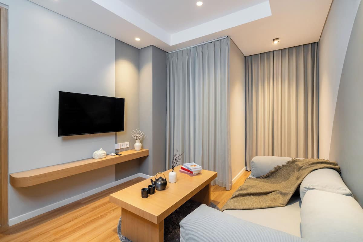 A modern living room with a flat-screen TV, wooden coffee table, sofa, and a large window with curtains.