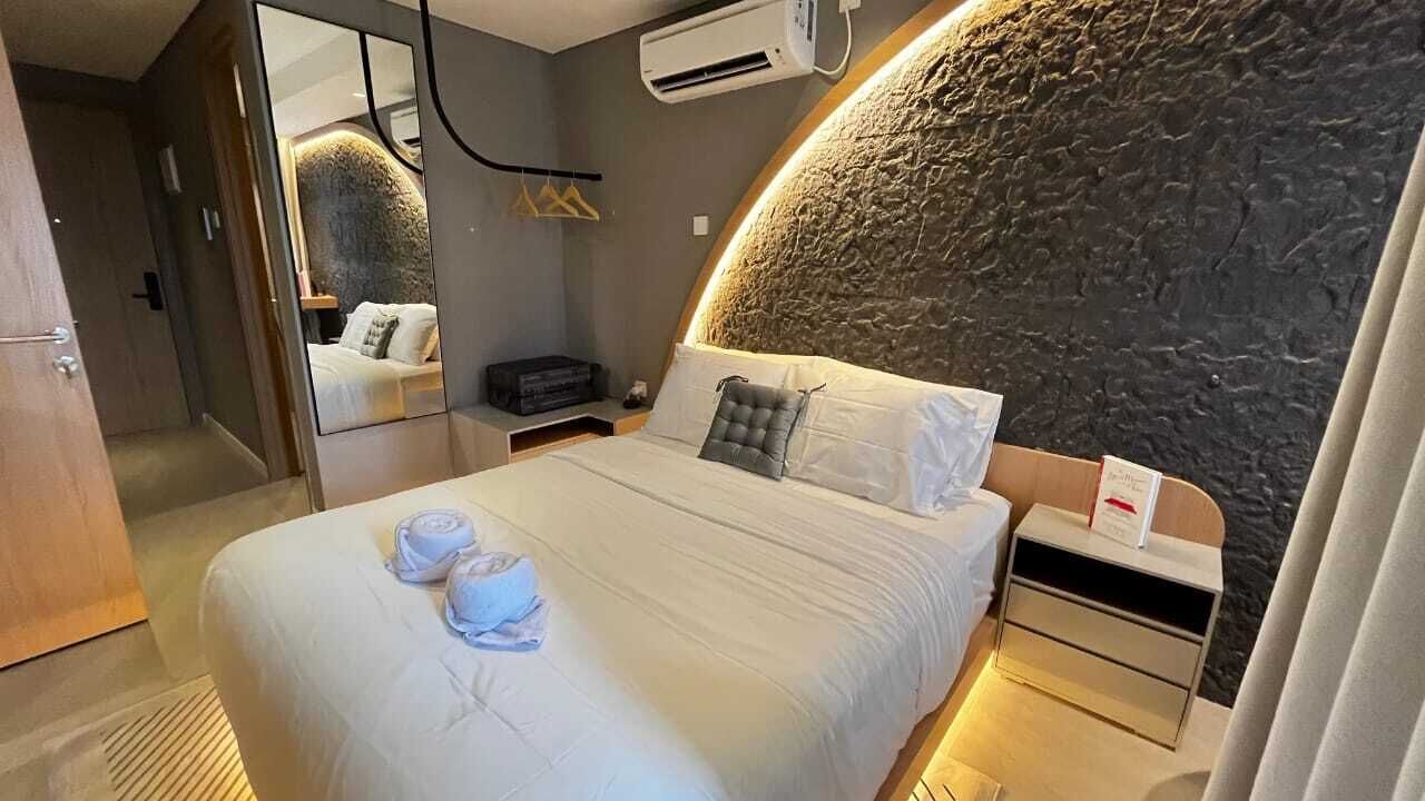 A modern hotel room with a curved stone wall, a large bed with white linens, a bedside table with a lamp, and a mirror reflecting the room.