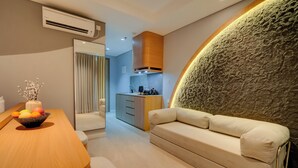 A modern living room with a sofa, a wooden table with fruit, and a kitchen in the background. - Morich Suite Antasari (Jakarta)