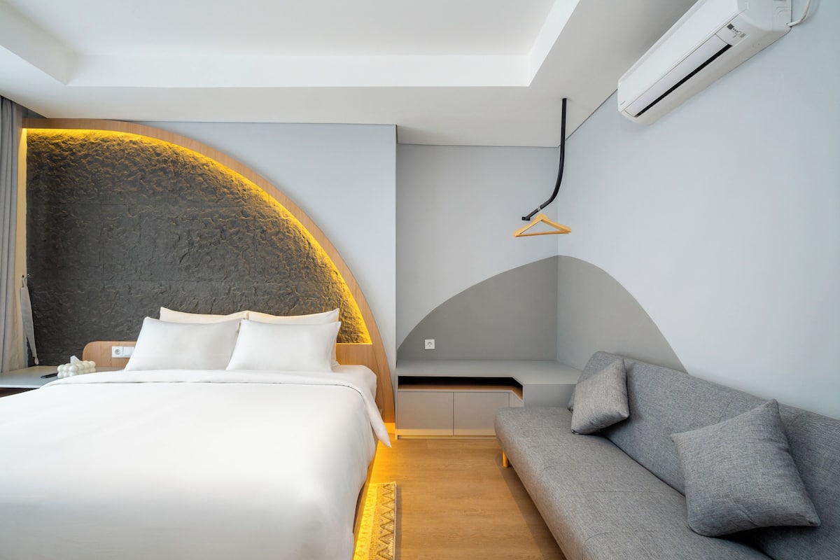 A modern hotel room with a curved stone wall, a bed with white linens, a grey sofa, and a built-in bench.