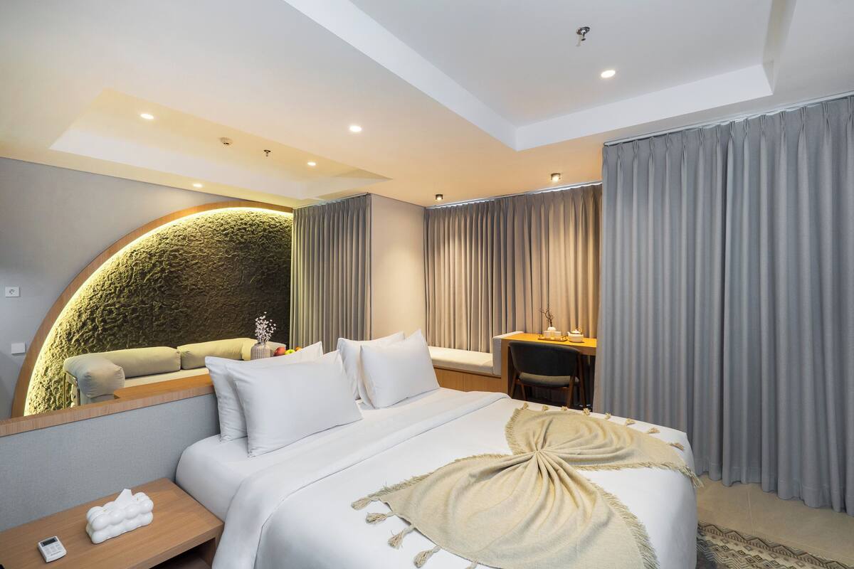 A modern hotel room with a bed, bedside table, desk, and a curved wall feature.