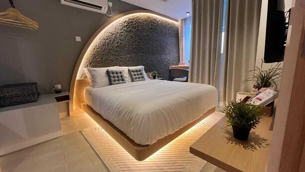 A modern bedroom with a large bed, a round stone headboard, a bedside table with a plant, and a small desk with a chair. - Morich Suite Antasari (Jakarta)