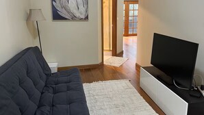 Un salon avec un canapé foncé, un téléviseur à écran plat posé sur un meuble blanc, une lampe et un tableau accroché au mur.