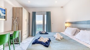 Une chambre d’hôtel avec un lit, un bureau, des chaises vertes, une vue sur la mer et un tableau au mur.