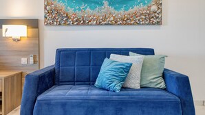 Un sofá azul con dos almohadas, una lámpara de pared y un cuadro de una playa.