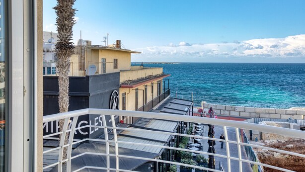 A balcony with a view of the sea, a building with a sign 'ACLES', and a palm tree. - Stylish Apartment in Bay Square 1 (St. Paul's Bay)