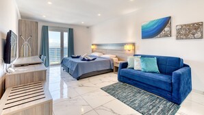 Quarto de hotel moderno com uma cama, um sofá azul, uma televisão e uma varanda com vista para o mar.