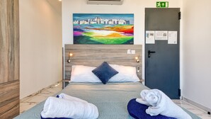 Habitación de hotel con cama, dos toallas, un cuadro de un paisaje urbano y una puerta con señal de salida de emergencia.