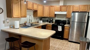 A modern kitchen with wooden cabinets, a stainless steel refrigerator, a microwave, and a stove. There are two bar stools and a trash can. - Urban Comfort with Northern Charm (Anchorage)