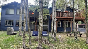 A two-story house with a deck, surrounded by trees and a grassy yard. - Urban Comfort with Northern Charm (Anchorage)