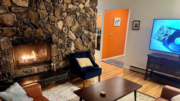 A living room with a stone fireplace, a blue armchair, a brown leather sofa, a wooden coffee table, and a television on a stand. - Urban Comfort with Northern Charm (Anchorage)