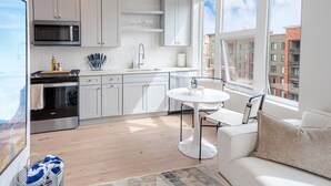 Una cocina moderna con armarios blancos, un microondas y una estufa. Hay un comedor con una mesa y sillas blancas, y un sofá con un cojín.