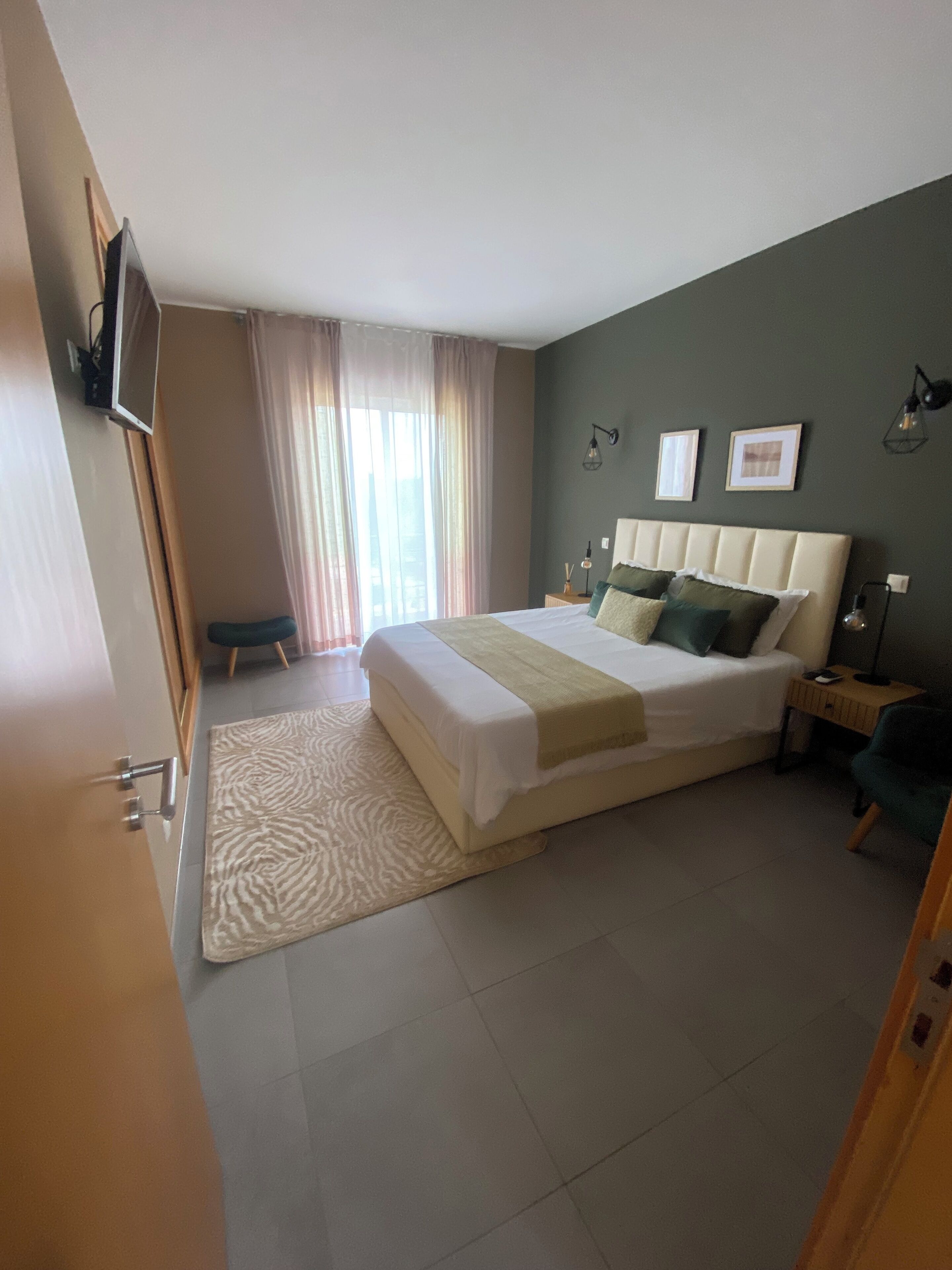 A bedroom with a large bed, a desk, a chair, a TV mounted on the wall, and a window with curtains.