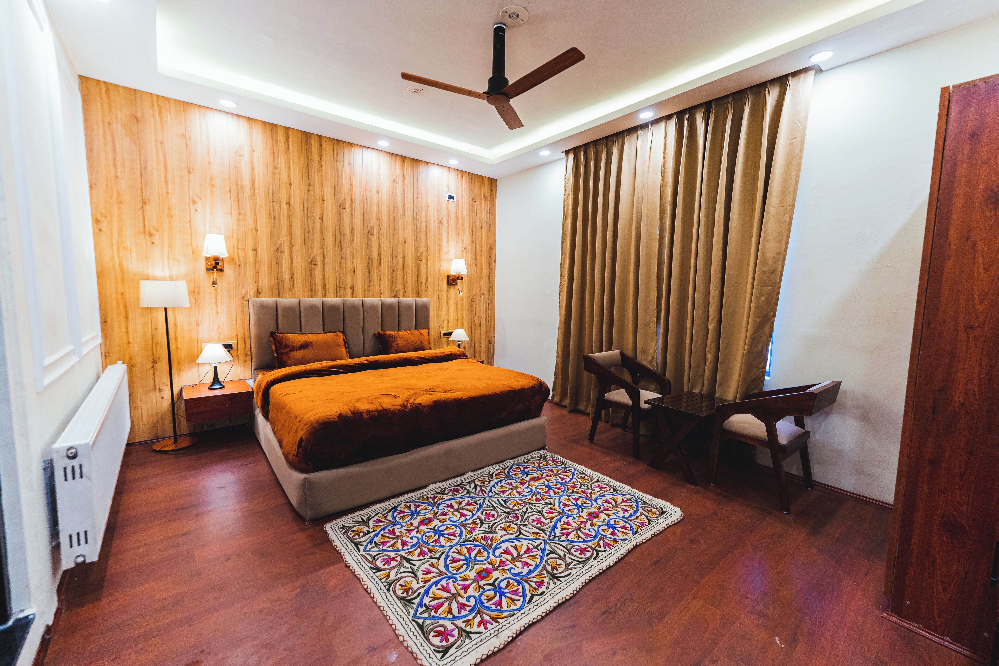 A bedroom with a wooden wall, a bed with an orange bedspread, a desk with a chair, and a patterned rug.