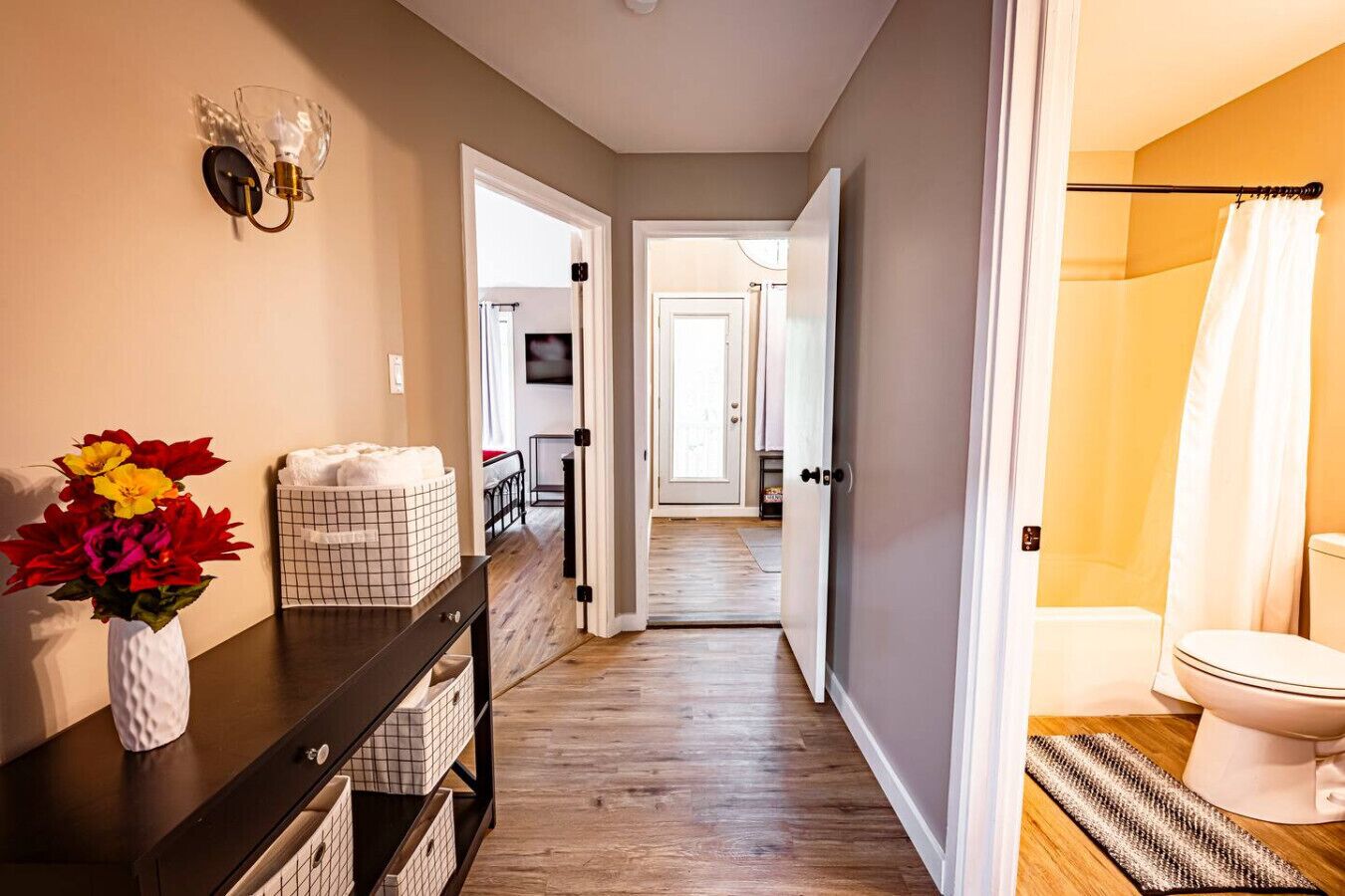 Un couloir doté d’un plancher en bois, une console noire avec des paniers de rangement, un vase avec des fleurs et une porte donnant sur une salle de bain équipée de toilettes et d’un rideau de douche.