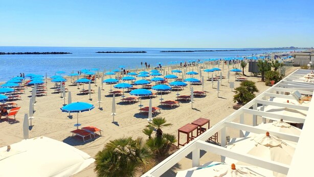 Une plage parsemée de nombreux parasols bleus et de chaises longues blanches, un ciel dégagé et une mer calme.