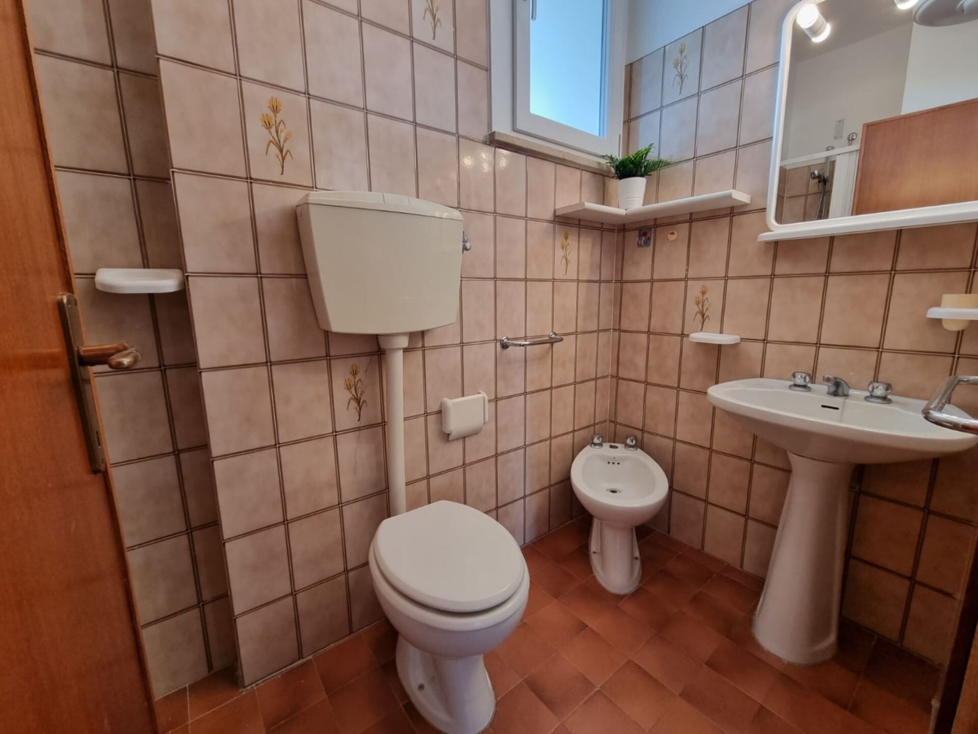 Ein Badezimmer mit Toilette, Bidet und Waschbecken, gefliesten Wänden und einem Fenster.