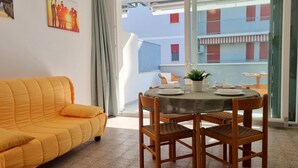 Une pièce avec un canapé jaune, une table à manger avec des chaises, et une vue sur un immeuble à travers une porte vitrée.