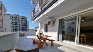 Un balcon avec une table et des bancs en bois, un canapé en osier et une porte-fenêtre coulissante donnant sur la cuisine.