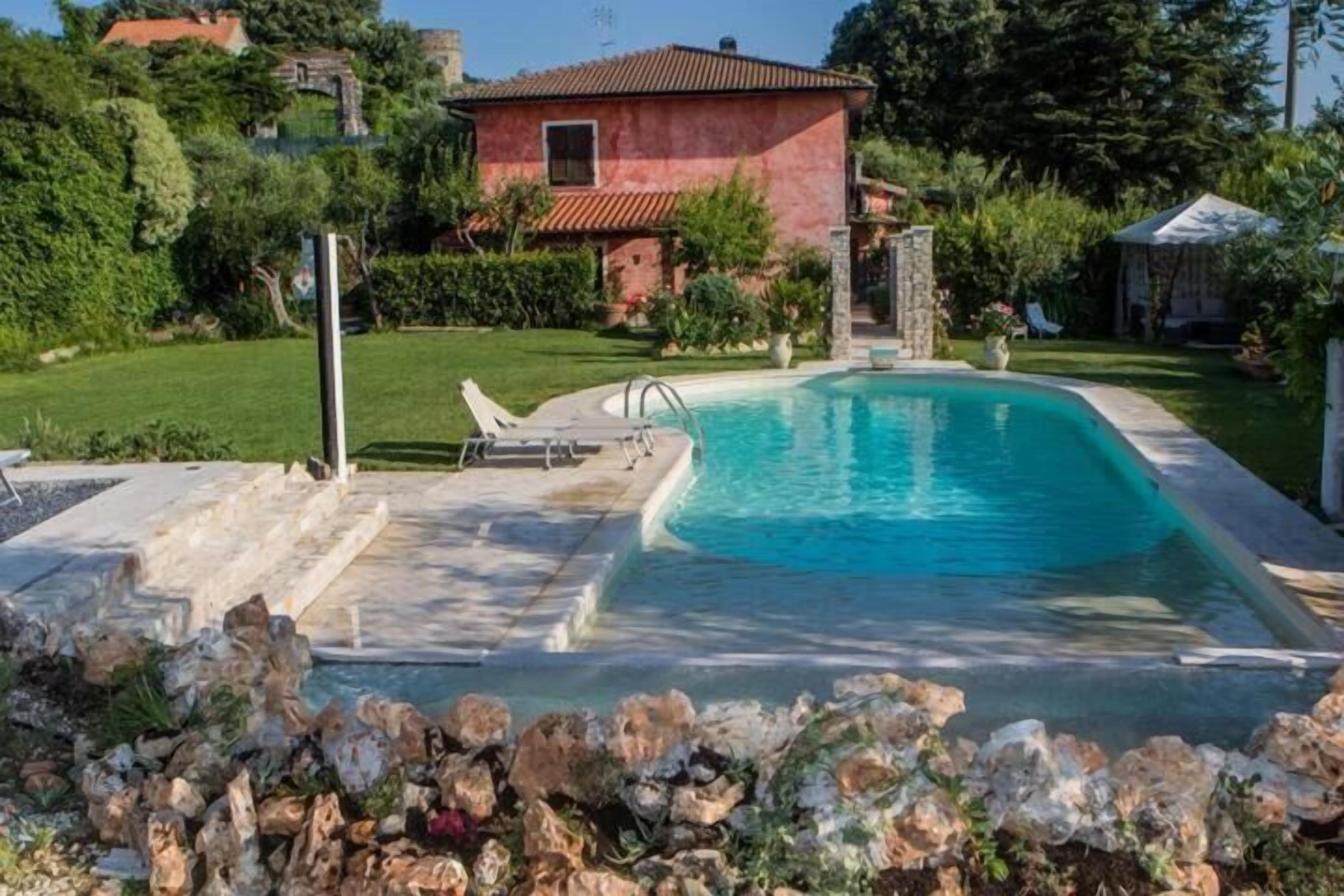 A swimming pool with a stone border, a stone patio, and a red-roofed house in the background.