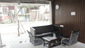 A reception area with a black leather sofa, two metal-framed chairs, a small table with a newspaper, and a clock on the wall. - Hotel Hare Krishna Vrindavan (Vrindavan)