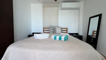 Uma cama bem arrumada com um edredom, duas toalhas enroladas, uma mesa de cabeceira, um espelho e um ventilador de teto.