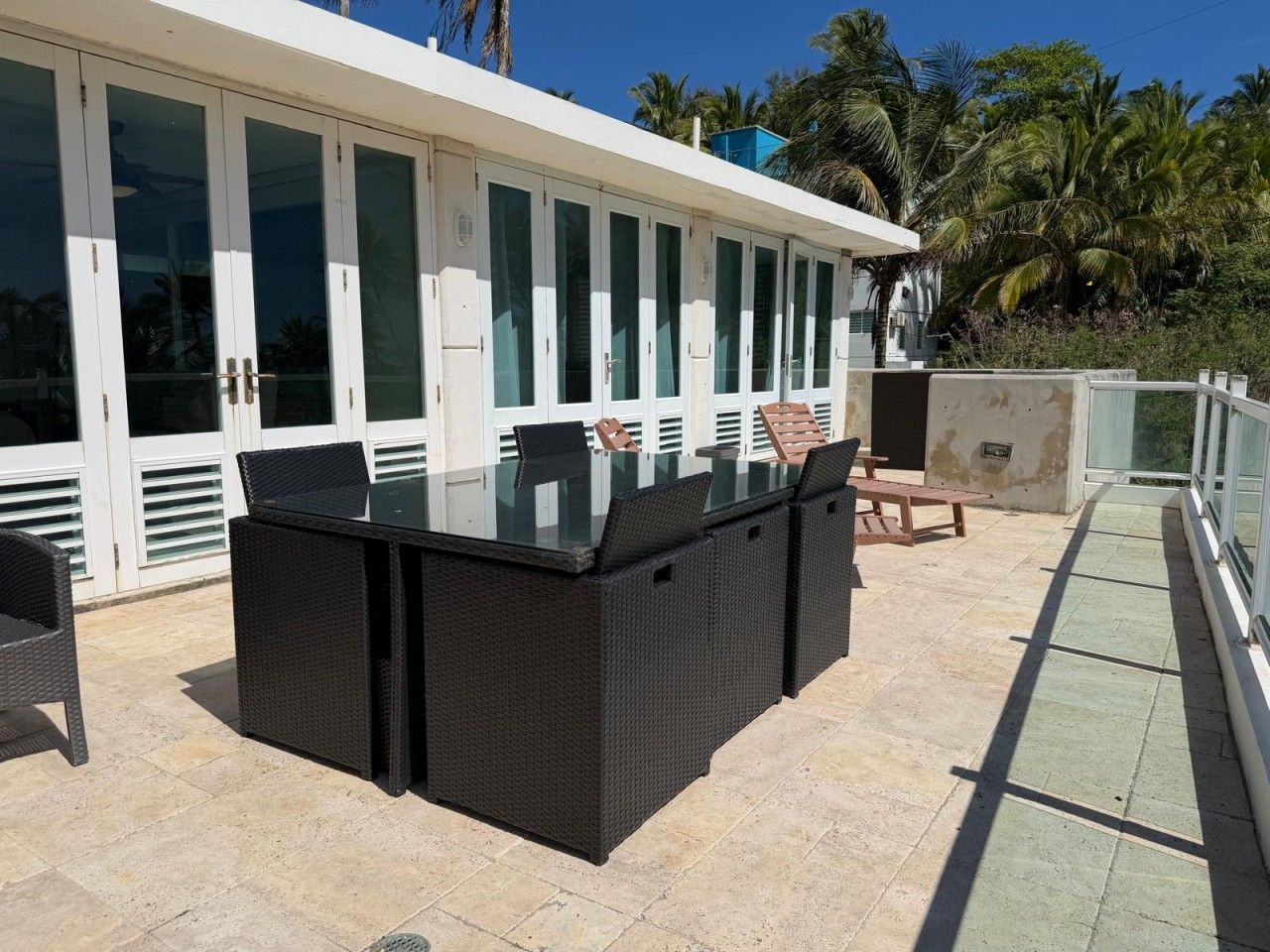 An outdoor patio with a glass-top table and wicker furniture, adjacent to a building with large windows and a balcony.