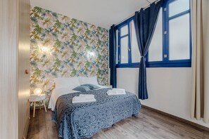 Un dormitorio con cama, mesita de noche, pared con estampado de hojas tropicales, ventana con cortinas azules y piso de madera.