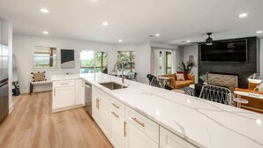 A modern kitchen with white cabinetry and a large island, a dining area with a sofa and chairs, and a flat-screen TV mounted on the wall.