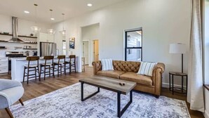 Un salon moderne avec un canapé en cuir marron, une table basse en bois et un tapis à motifs. On aperçoit à l’arrière-plan une cuisine avec des meubles blancs et des appareils électroménagers en acier inoxydable.