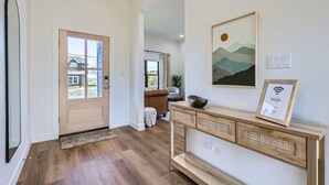 Un hall d’entrée moderne avec un plancher en bois, une porte beige et une console sur laquelle sont posés un tableau encadré et un panneau indiquant la présence d’un réseau Wi-Fi.