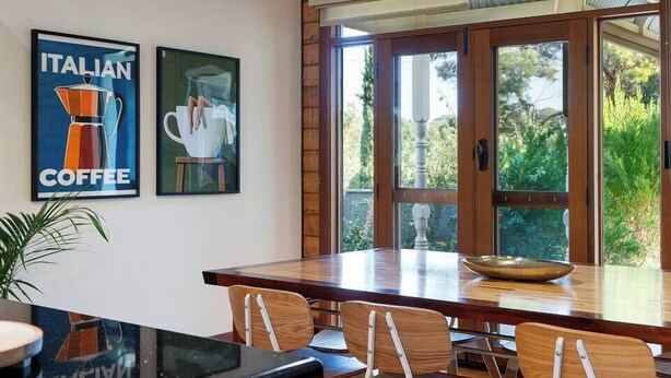 Un coin repas avec une table et des chaises en bois, de grandes fenêtres donnant sur un paysage verdoyant, et deux tableaux encadrés accrochés au mur.