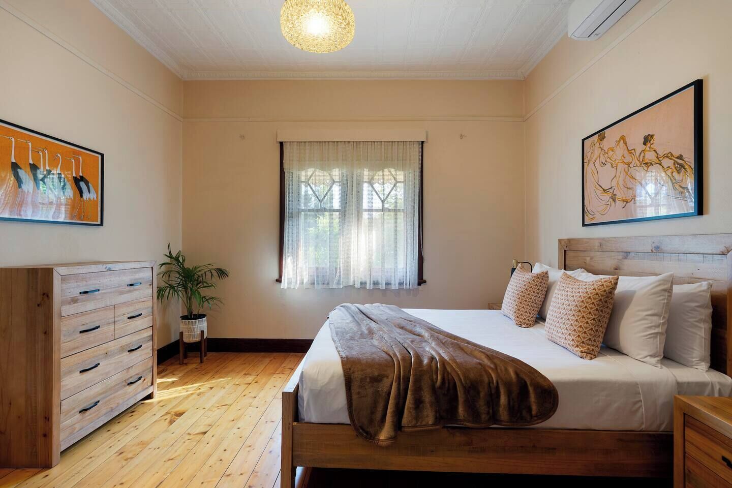 Quarto com cama de madeira, cômoda, janela com cortinas leves e duas pinturas nas paredes.