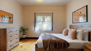 Quarto com cama de madeira, cômoda, janela com cortinas leves e duas pinturas nas paredes.