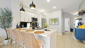 A modern kitchen with white cabinets, a central island with bar stools, a hanging light fixture, and a potted plant.