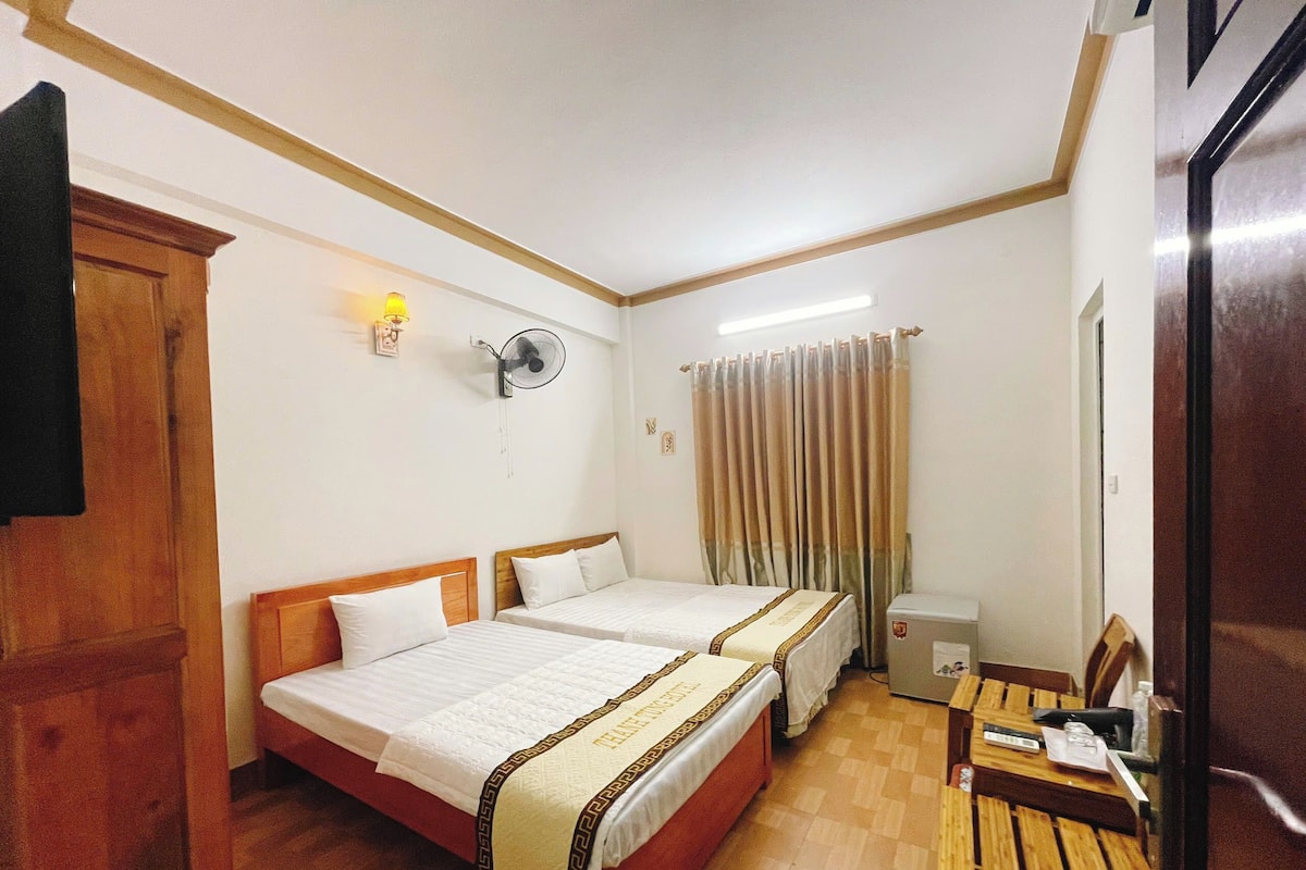 Ein Hotelzimmer mit zwei Betten, einem Holztisch, einem Ventilator und einem Vorhang.