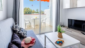 Un salon moderne avec un canapé, un téléviseur à écran plat, une petite table avec une plante et un balcon équipé d’une table et de chaises.
