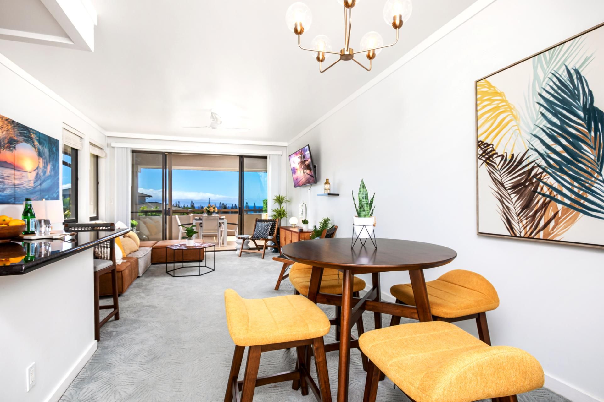 A modern living room with a large window, a dining area with yellow chairs, and a TV mounted on the wall.