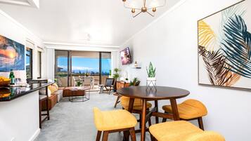A modern living room with a large window, a dining area with yellow chairs, and a TV mounted on the wall.